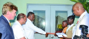 Acting President Samuel Hinds cuts the ribbon to declare open the Supply Chain Management Complex as Health Minister, Dr Bheri Ramsaran; U.S. Ambassador to Guyana, D Brent Hardt; Junior Finance Minister Juan Edghill and schoolchildren look on